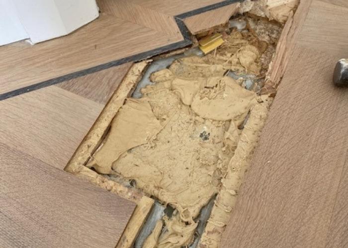 Close-up van een houten vloer waar een beschadigd gedeelte is verwijderd voor reparatie. De onderliggende structuur en oude materialen zijn zichtbaar terwijl Vloerenonderhoud Nederland bezig is met het herstellen van de vloer. Gereedschap ligt klaar voor gebruik tijdens het reparatieproces.
