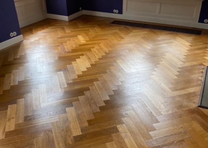 Een prachtig afgewerkte visgraat houten vloer in een woonruimte, met een warme, glanzende afwerking die het vakmanschap van Vloerenonderhoud Nederland weerspiegelt. De vloer ligt in een kamer met paarse muren en witte plinten.