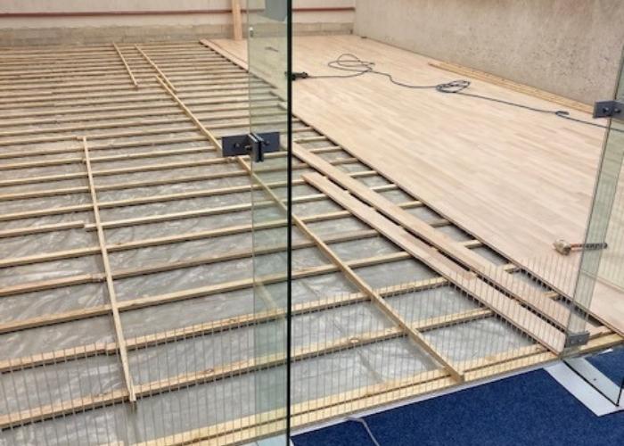 Een gedeeltelijk geïnstalleerde houten vloer in een ruimte, waarbij de onderliggende houten frameconstructie zichtbaar is. De vloerplanken worden zorgvuldig geplaatst door Vloerenonderhoud Nederland